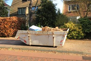 Ein weißer Müllcontainer voller Pappkartons und anderem Abfall, geparkt auf einer Straße vor Häusern und Laub.